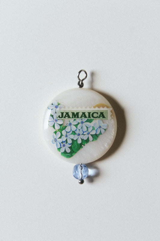 A round pendant charm featuring a white background with a green 'JAMAICA' stamp and blue-and-white floral artwork, hanging from a small silver loop with a light blue bead below, photographed on a plain light background.